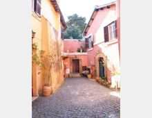 Cortile interno acciottolato tra edifici dai muri color ocra e rosa, con finestre a persiane e portoni ad arco in mattoni. Lungo le pareti ci sono vasi e fioriere con piante, e sul fondo un ingresso con cancello sotto una piccola tettoia.