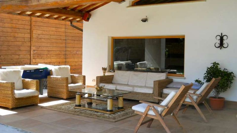 Veranda coperta con travi in legno a vista e pavimento piastrellato, arredata con divani e poltrone in vimini con cuscini chiari, tavolino in vetro e sedie reclinabili in legno. Sullo sfondo una parete bianca con grande finestra, pannelli in legno laterali e un vaso con pianta.