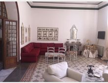 Salotto in stile classico con pavimento in piastrelle decorate, grande divano angolare bordeaux e sedie bianche attorno a un tavolino. Sulla parete si vedono stampe incorniciate e, a destra, un camino con specchiera ornata; a sinistra una porta ad arco in legno con vetri.