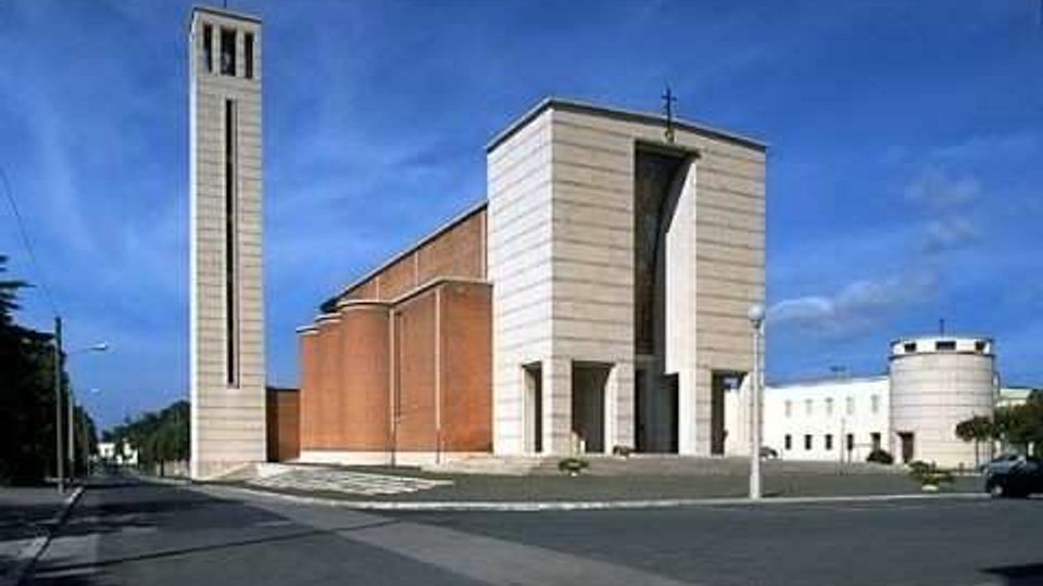 Chiesa moderna con facciata in pietra chiara e navata laterale in mattoni, affiancata da un alto campanile rettangolare con croce in cima. Davanti si apre un’ampia piazza asfaltata con strada e lampioni, con un edificio bianco sullo sfondo sotto un cielo azzurro.
