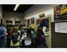 Interno di un cinema con folla in coda davanti alla biglietteria incassata nel muro, con display luminoso sopra lo sportello. Sulle pareti sono affissi poster di film e a destra è visibile una piantina dell’edificio su pannello.