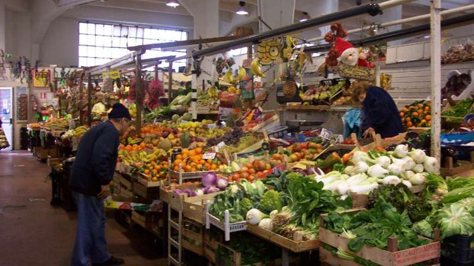Interno di un mercato coperto con grandi finestre e soffitto alto, con una lunga fila di banchi ortofrutticoli in cassette di legno colmi di frutta e verdura. Strutture metalliche sopra i banchi sostengono prodotti appesi e decorazioni.
