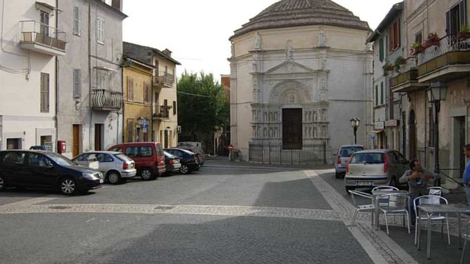 Piazza lastricata in un centro storico, con edifici residenziali ai lati e auto parcheggiate lungo la strada. Sullo sfondo si trova una chiesa in pietra con facciata decorata e grande portale, coperta da una cupola ottagonale con tetto in tegole.