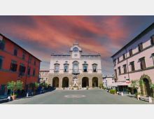 Piazza cittadina con al centro un edificio storico in stile classico, facciata bianca con portico ad archi al piano terra e balconi al piano superiore. La piazza è circondata da palazzi colorati e presenta una fontana centrale e vasi con piccoli alberi lungo i lati.