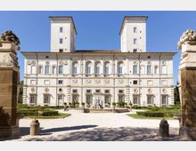Facciata di un grande palazzo storico in stile rinascimentale, con due torri quadrate sul retro, finestre ad arco e decorazioni in stucco su più livelli. Davanti si apre un giardino formale con siepi geometriche, vialetti in ghiaia e una fontana centrale.