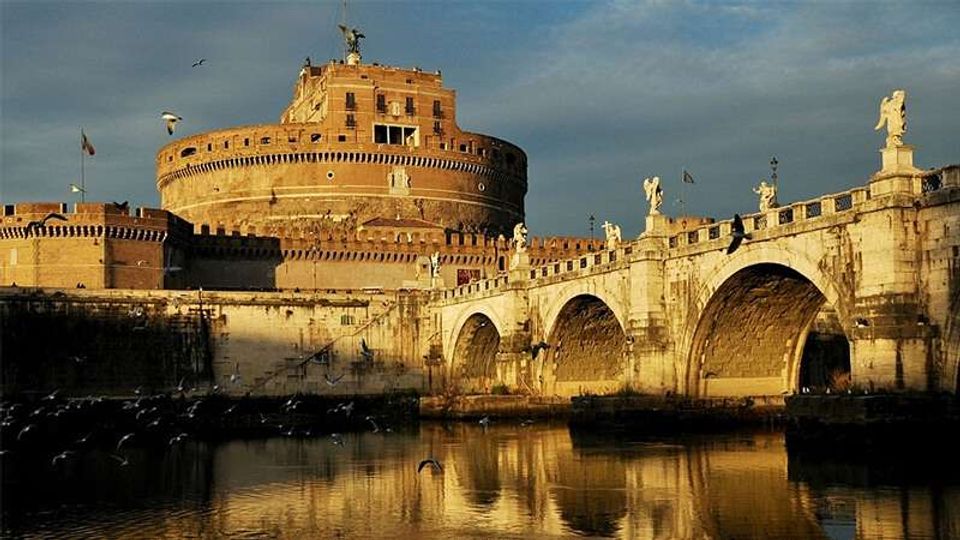 Veduta di Castel Sant’Angelo con la sua struttura cilindrica in muratura e, in primo piano, il Ponte Sant’Angelo in pietra con arcate e statue lungo il parapetto. Il ponte attraversa il fiume, che riflette le pareti illuminate e il cielo.