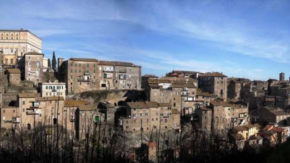 Panorama di un borgo collinare con case in pietra addossate su più livelli e tetti in coppi, disposto lungo il pendio. In alto spicca un grande edificio storico con facciata chiara, circondato da altri fabbricati e mura, sotto un cielo azzurro.