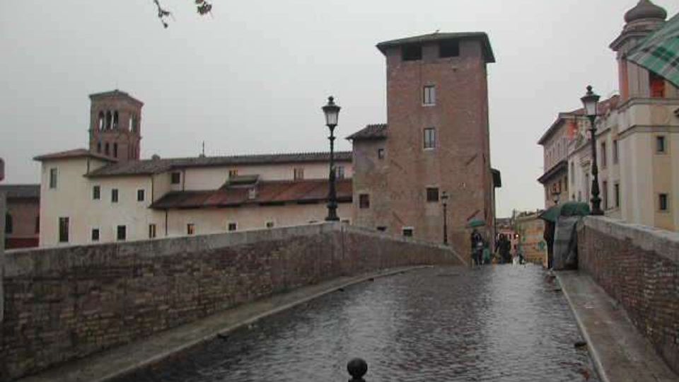 Ponte pedonale in pietra con pavimentazione in ciottoli e parapetti in muratura, che conduce verso edifici storici con torri in mattoni. Lungo il ponte si vedono lampioni e, sullo sfondo, un complesso di case e strutture d’epoca sotto un cielo grigio.