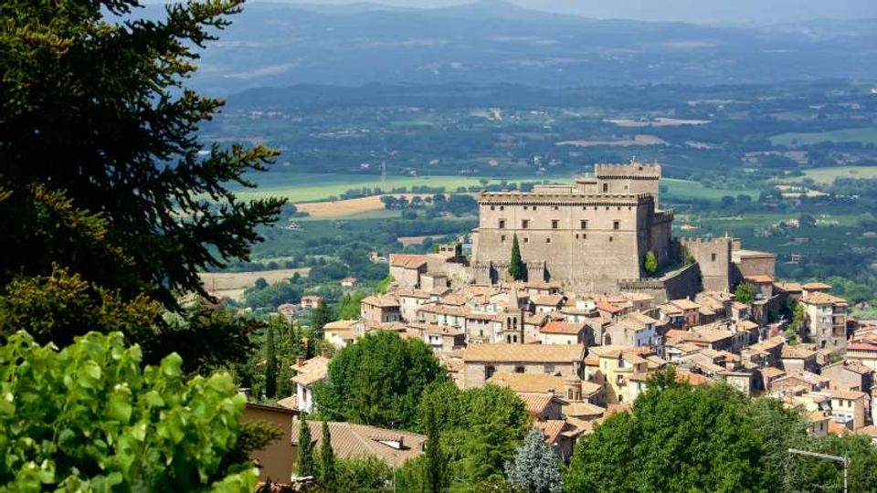 Veduta panoramica di un borgo collinare con case in pietra dai tetti in coppi, dominato da un grande castello-fortezza rettangolare con torri. Sullo sfondo si estende una valle con campi e boschi, con colline e montagne lontane.