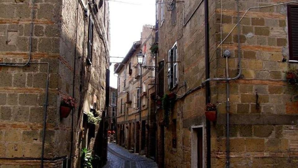 Stretta via acciottolata tra alti edifici in pietra, con persiane alle finestre, vasi di fiori alle pareti e lampioni lungo il percorso. Cavi e tubazioni corrono sulle facciate, mentre la strada curva e si restringe verso il fondo.