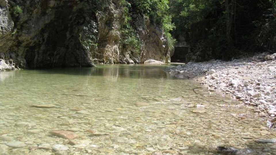 Ruscello dalle acque limpide e poco profonde in una gola rocciosa, con fondale di ciottoli visibili e una riva ghiaiosa. Pareti di roccia e fitta vegetazione alberata incorniciano il corso d’acqua.
