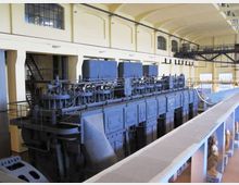 Grande sala industriale con soffitto alto ad arcate e finestre ad arco, vista da una passerella sopraelevata con ringhiere. Al centro si trova un enorme macchinario blu con tubazioni e pannelli metallici, tipico di una centrale o impianto storico.