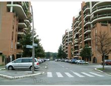 Strada residenziale ampia con attraversamento pedonale, auto parcheggiate ai lati e alberi lungo i marciapiedi. Su entrambi i lati si affacciano alti condomini in mattoni con balconi curvi e ringhiere bianche, con un palo segnaletico all’incrocio in primo piano.