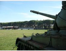Campo erboso all’aperto con alberi sullo sfondo e una tribuna o edificio basso in lontananza. In primo piano si vede parzialmente un carro armato con cannone rivolto verso sinistra.