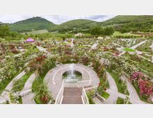 Giardino paesaggistico con aiuole fiorite e pergolati, organizzato in vialetti curvi attorno a una fontana circolare centrale con getto d’acqua. Sullo sfondo si vedono colline verdi e campi coltivati, con alberi e piccoli edifici sparsi nel paesaggio.