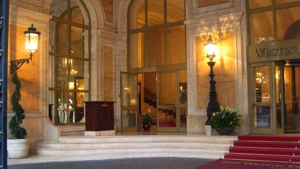 Ingresso di un hotel con facciata in pietra, grandi finestre e porte ad arco in vetro e gradini in marmo. Due lampioni a parete illuminano il portico, con tappeto rosso sulla scalinata e vasi con piante ai lati.