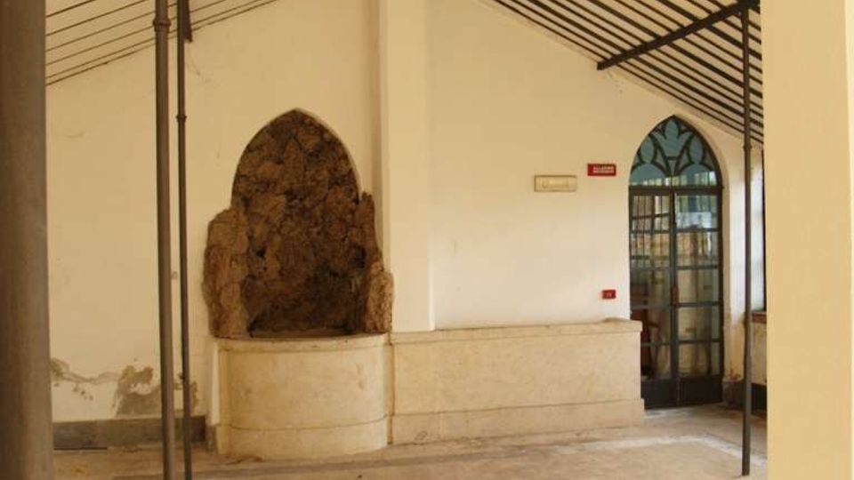 Interno di un ambiente in stile industriale con pareti bianche e soffitto inclinato a pannelli e travi metalliche, sostenuto da sottili colonne scure. Sul lato sinistro c’è una nicchia ad arco con roccia a vista sopra una base in pietra, mentre a destra si vede una porta ad arco vetrata con inferriate.