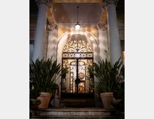 Ingresso di un edificio classico con due alte colonne e un portale ad arco, chiuso da un cancello in ferro battuto decorato e illuminato da una lanterna sospesa. Ai lati della scalinata ci sono grandi vasi con piante, con l’interno visibile in controluce oltre il cancello.