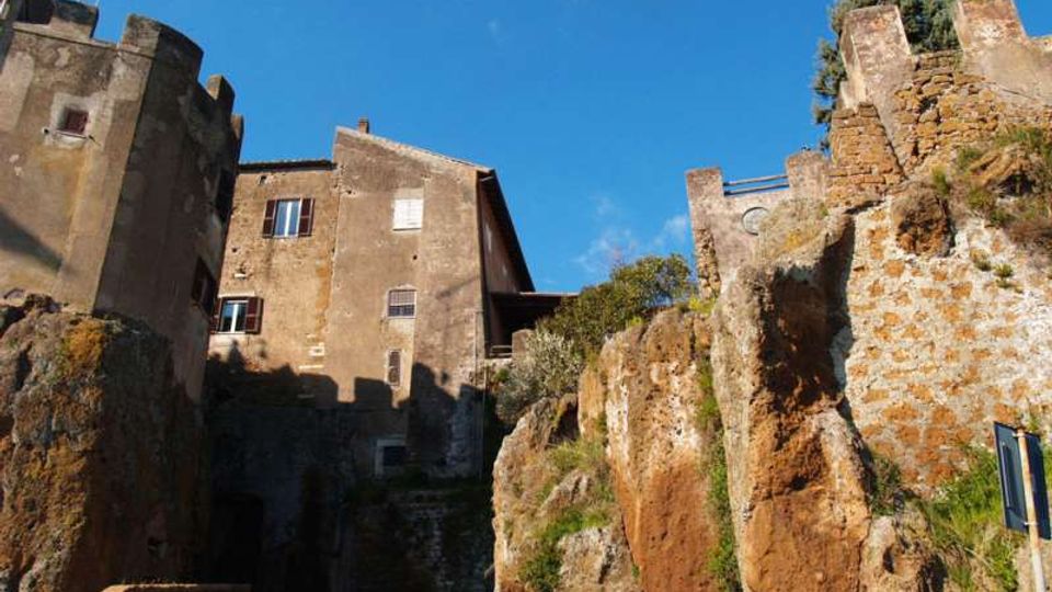 Case in pietra e intonaco di un borgo storico costruite su una parete di roccia tufacea, con resti di mura e vegetazione sulla scarpata. Strada asfaltata in basso e cielo sereno sullo sfondo.