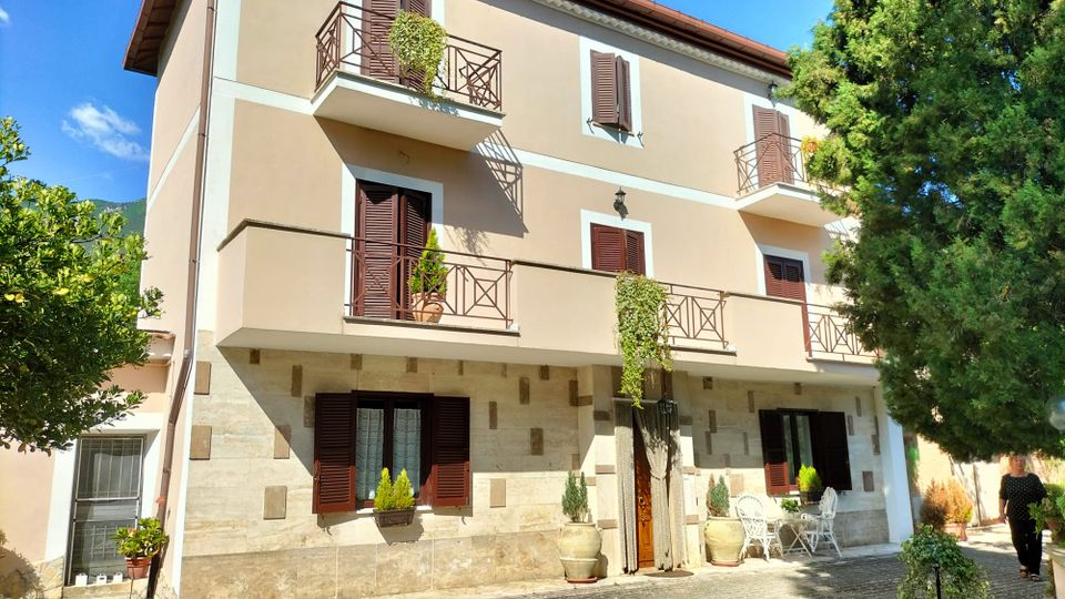 Facciata di una palazzina residenziale a tre piani con intonaco beige e base in pietra, persiane in legno scuro e balconi con ringhiere metalliche e piante ricadenti. Davanti c’è un cortile pavimentato con vasi e un piccolo set da esterno, circondato da alberi sotto un cielo azzurro.