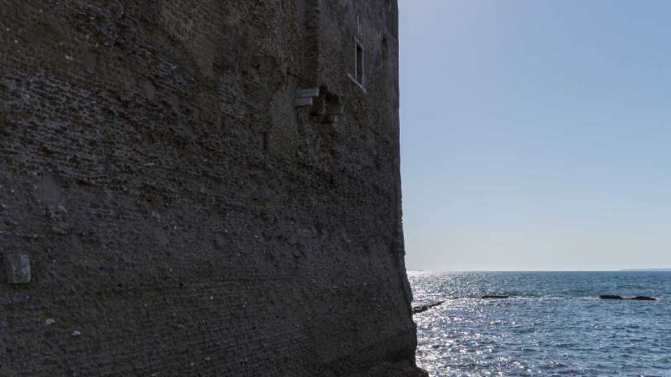 Vista laterale di una massiccia fortificazione in pietra con merlature, affacciata direttamente sul mare. L’acqua lambisce la base del muro mentre il sole alto illumina l’orizzonte marino.