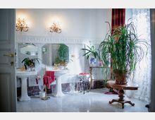 Bagno ampio e luminoso con pavimento in marmo, due lavabi su colonna con rubinetteria dorata e specchi a parete, illuminati da applique classiche. Vicino a una grande finestra con tende e drappeggi rossi, un tavolino in legno sostiene una grande pianta in vaso.
