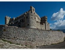Castello medievale in pietra con merlature e torri, visto dal basso dietro un’alta cinta muraria in pietra. Sullo sfondo cielo azzurro con nuvole e colline lontane.