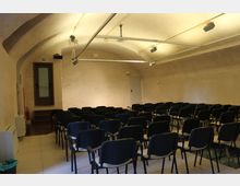 Sala conferenze sotterranea con soffitto a volta intonacato e illuminazione a binari sospesi. File di sedie nere disposte verso il fondo della stanza, con una porta laterale su un piccolo rialzo e pareti color crema.