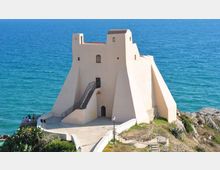 Torre costiera fortificata intonacata di bianco, con mura inclinate e scala esterna in metallo, costruita su uno sperone roccioso. Sullo sfondo si vede il mare aperto, con un piccolo piazzale e un sentiero di accesso davanti all’edificio.