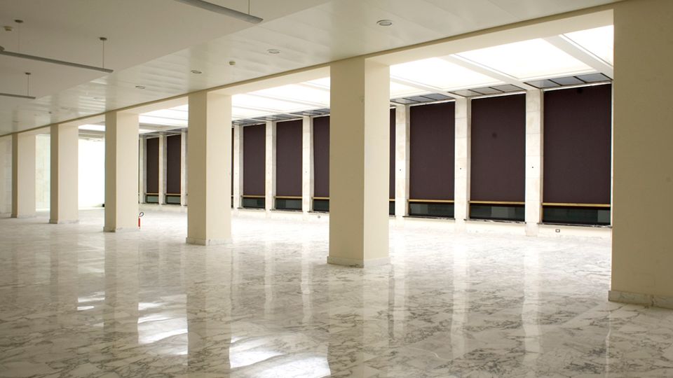 Ampio spazio interno vuoto, simile a una sala o open space, con colonne rettangolari in fila, pavimento in marmo lucido e soffitto con pannelli e illuminazione incassata. Lungo un lato si vedono grandi finestre schermate da pannelli scuri e lucernari che lasciano entrare luce naturale.