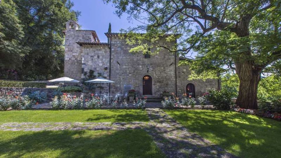 Edificio storico in pietra con facciata semplice, piccolo campanile e ingresso ad arco, affacciato su un giardino curato con prato, aiuole fiorite e tavoli all’aperto con ombrelloni. Grandi alberi e vegetazione fitta circondano l’area, con un sentiero che attraversa il prato.