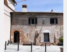 Facciata di un edificio in muratura intonacata e parzialmente scrostata, con tetto in coppi, comignolo e finestre con scuri scuri. Davanti si vede una piccola piazza lastricata con panchine in legno, paletti metallici e un lampione a parete.