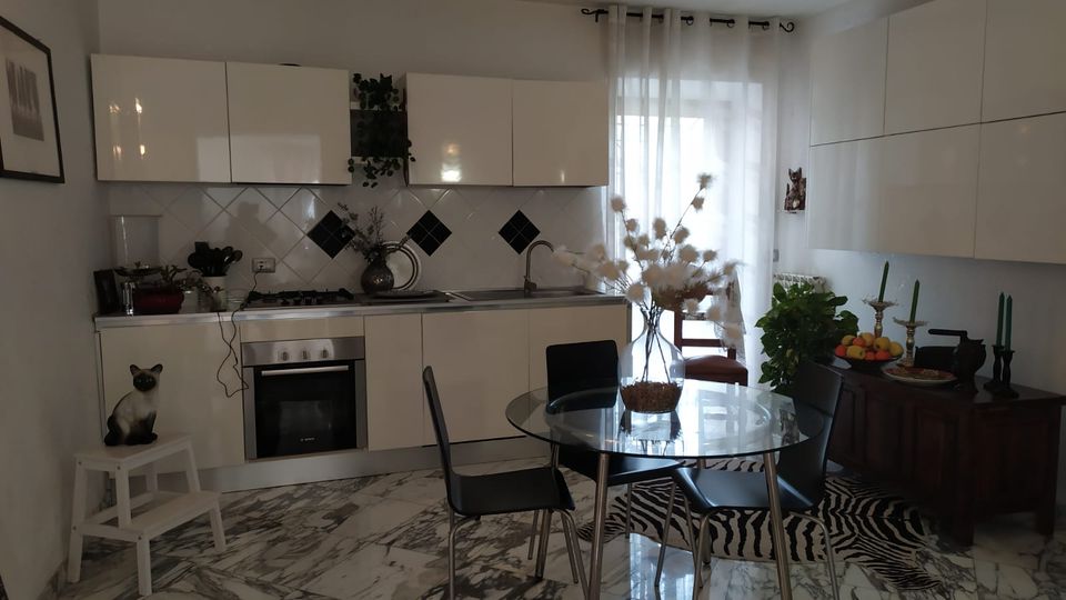 Cucina moderna con pensili bianchi lucidi e piano di lavoro lineare con fornelli e forno; parete piastrellata bianca con inserti a rombo neri. Al centro c’è un tavolo rotondo in vetro con sedie nere, pavimento effetto marmo e una grande finestra con tende leggere accanto a una credenza.