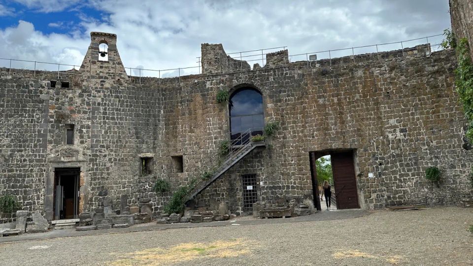 Cortile interno di una fortezza in pietra con alte mura merlate, un piccolo campanile e un arco sopraelevato raggiunto da una scalinata esterna. Sul lato destro si vede un grande portale aperto e, in alto, un camminamento con ringhiera sotto un cielo nuvoloso.