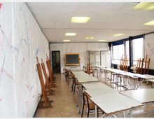 Aula scolastica adibita a laboratorio d’arte con lunghi tavoli e sedie in fila, cavalletti di legno appoggiati alle pareti e una lavagna in fondo. Grandi finestre su un lato illuminano l’ambiente, con pareti segnate da schizzi e un soffitto con pannelli luminosi.