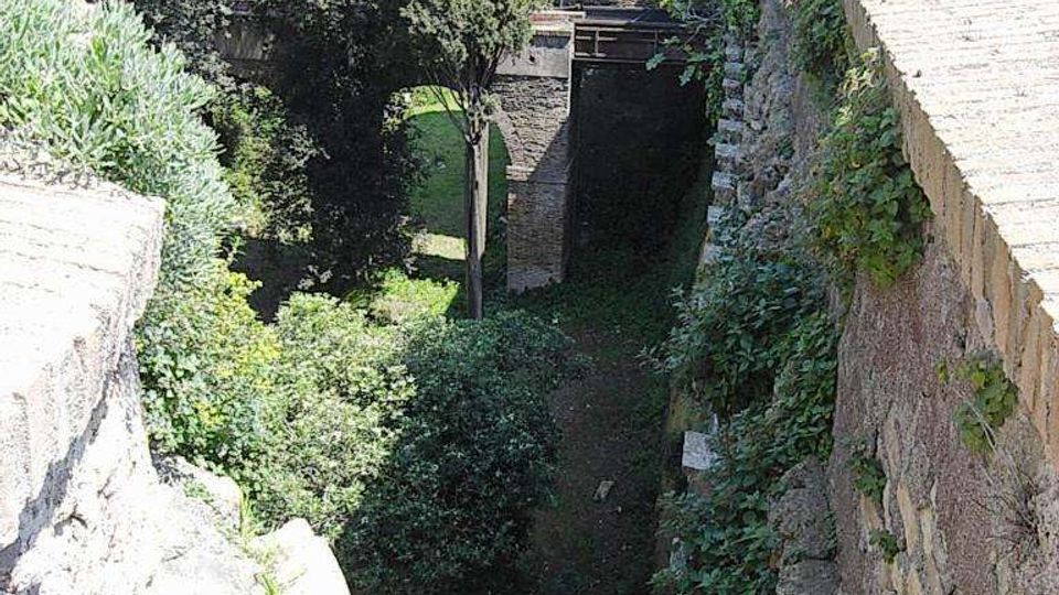Veduta dall’alto di un fossato lungo mura in pietra e mattoni, con pareti parzialmente coperte di edera. In basso si vede vegetazione fitta con alberi e un piccolo passaggio ad arco sotto una struttura muraria.