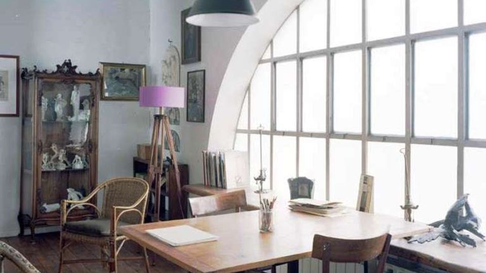 Interno di uno studio con grande finestra ad arco a riquadri che illumina un tavolo da lavoro in legno. Nella stanza si vedono pavimento in assi di legno, sedie in legno e vimini, una lampada a sospensione, una lampada da terra, quadri alle pareti e una vetrina con oggetti.