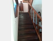 Scala interna in legno scuro con gradini lucidi e ringhiera intagliata, in un corridoio con pareti bianche. In cima alla rampa si vede un piccolo mobile a cassetti e un quadro appeso, con una finestra laterale che illumina lo spazio.