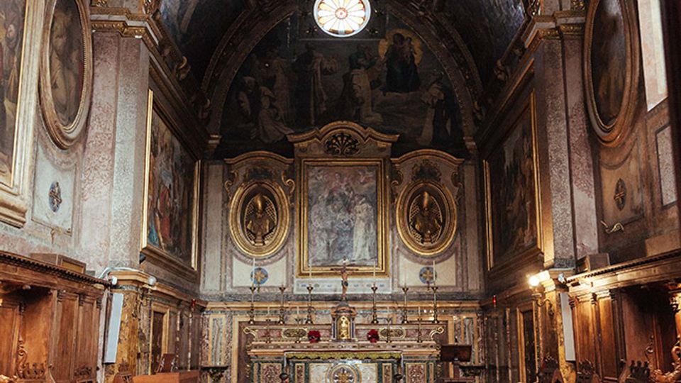 Interno di una cappella o piccola chiesa con navata unica, pavimento a scacchi e file di sedie in legno rivolte verso l’altare decorato. Volta a botte con affreschi e archi ornamentali, pareti con pannellature lignee e dipinti, e rosone circolare sopra il presbiterio.