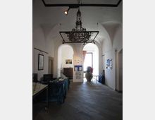 Interno di una sala espositiva con soffitto a volte bianche e grande lampadario in ferro battuto sospeso. Pavimento in pietra grigia, scrivania con computer a sinistra e pannelli espositivi con una grande finestra luminosa sul fondo.