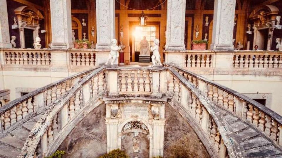 Cortile interno di un palazzo storico con doppia scalinata in pietra e balaustre, che converge verso una terrazza centrale. Al livello superiore si vedono colonne, nicchie con statue e un portale illuminato sullo sfondo.