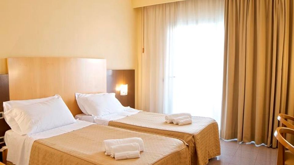 Camera d’albergo con due letti singoli affiancati, testiera in legno e biancheria chiara con asciugamani piegati. Ampia portafinestra con tende beige che lascia entrare luce naturale su pavimento piastrellato.