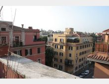 Veduta urbana da una terrazza con muretto in primo piano, affacciata su edifici residenziali di più piani con facciate colorate e tetti in coppi. Sullo sfondo si vede lo skyline della città con altri palazzi e alberi lungo la strada sottostante.