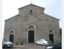 Facciata in pietra di una chiesa in stile romanico, con frontone triangolare sormontato da una croce, una piccola finestra ad arco e tre ingressi con portali ad arco. Davanti si vede una piazza pavimentata con alcune auto parcheggiate e grandi fioriere.