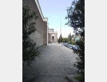 Ampio marciapiede in pietra che costeggia un grande edificio moderno rivestito in blocchi, con ingresso arretrato e alti pennoni. Sul lato destro corre una strada con auto parcheggiate, alberi e cespugli che incorniciano la vista.