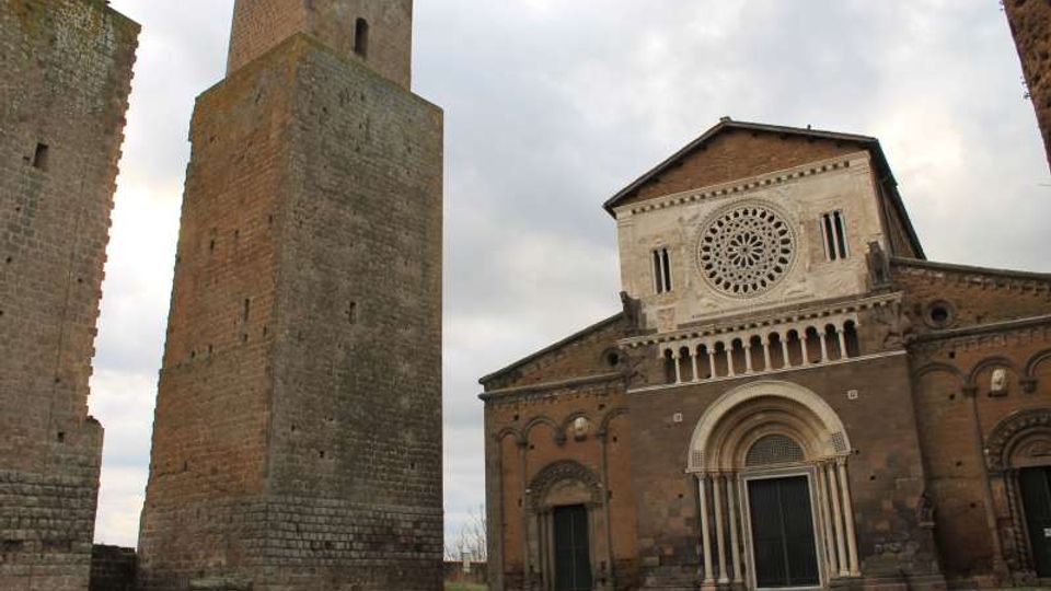 Facciata di una chiesa romanica in pietra con grande rosone centrale e portale ad arco, affiancata da un’alta torre medievale in muratura. Davanti si estende un prato, con cielo nuvoloso sullo sfondo.
