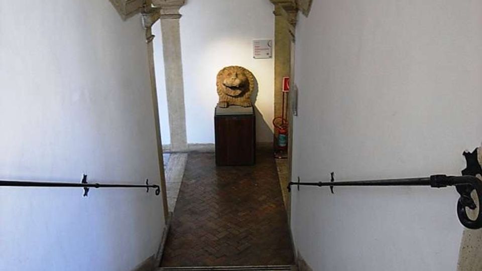 Scala interna in pietra che scende verso un corridoio con pavimento in mattoni e pareti bianche, dotata di corrimano in ferro ai lati. In fondo si vede un arco in pietra e una scultura di leone su piedistallo, illuminata da una luce a parete.