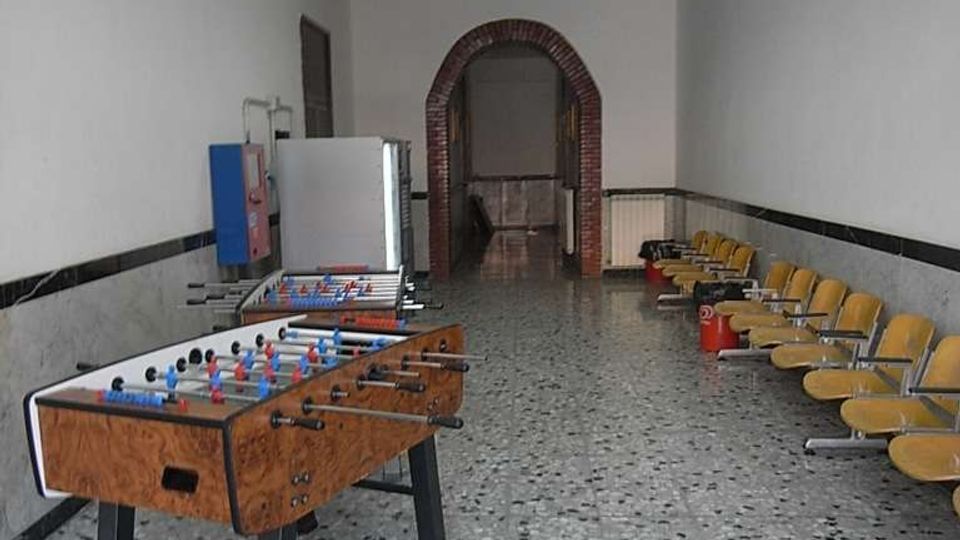 Sala ricreativa rettangolare con pavimento in graniglia e soffitto a pannelli, con due biliardini al centro e una fila di sedie gialle lungo la parete destra. In fondo si vede un arco in mattoni che conduce a un’altra stanza, con pareti bianche rivestite a mezza altezza.