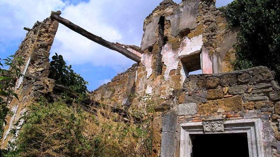 Rovine di un edificio in pietra con muri parzialmente crollati e intonaco consumato, con una trave in legno inclinata tra due pareti. In primo piano si vede un’apertura con cornice in pietra scolpita, circondata da vegetazione e sotto un cielo parzialmente nuvoloso.