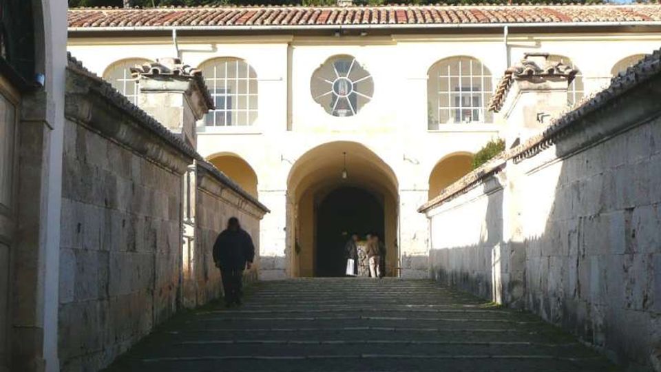 Scalinata in pietra tra due alti muri che conduce a un edificio chiaro con portico ad archi, finestre ad arco e rosone centrale sotto un tetto in coppi. Sullo sfondo si vedono alberi e un pendio collinare.
